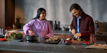 Woman in pink sweater cooking on hob next to man in red jacket cutting vegetable
