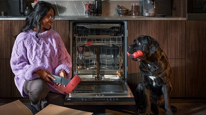 Kvinde pakker reservedele op til opvaskemaskine mens hun smiler til en sort hund som har hundelegetøj i munden