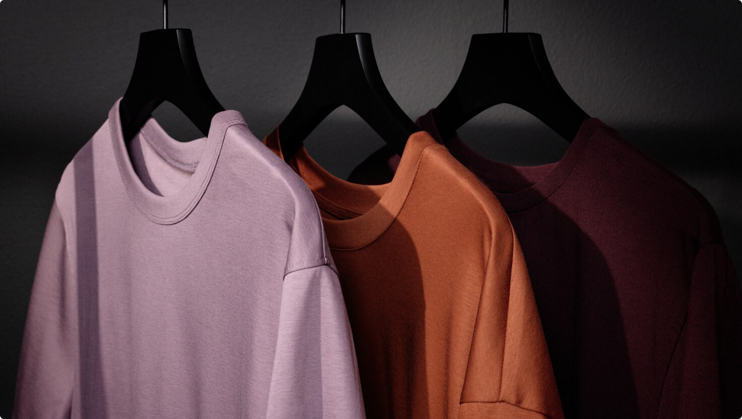 En lilla, en orange og en vinrød T-shirt hænger på hver sin sorte bøjle.